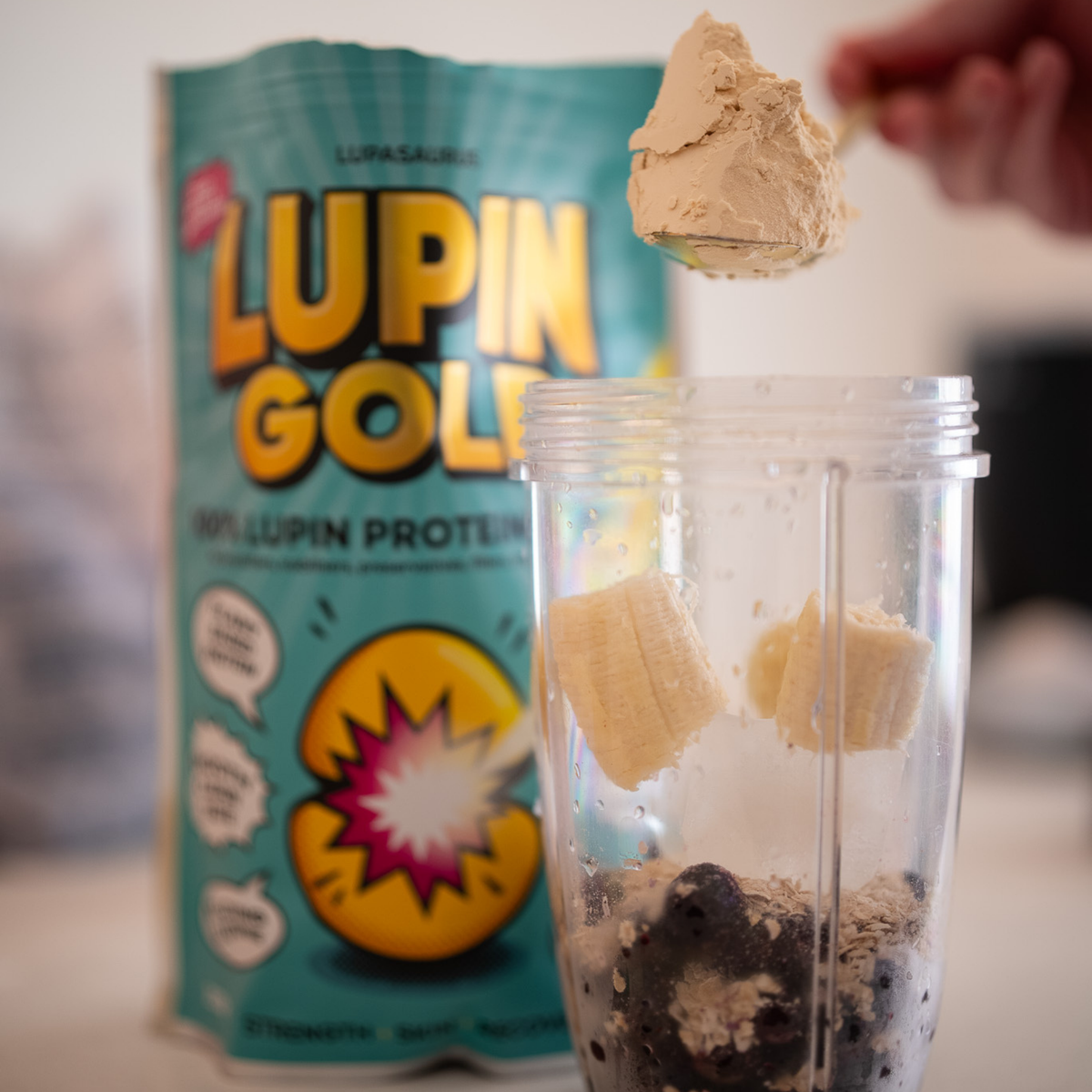 Scoop of unflavoured lupin protein powder being tipped into a blender cup of banana, blueberries and oats beside a Lupin Gold pack