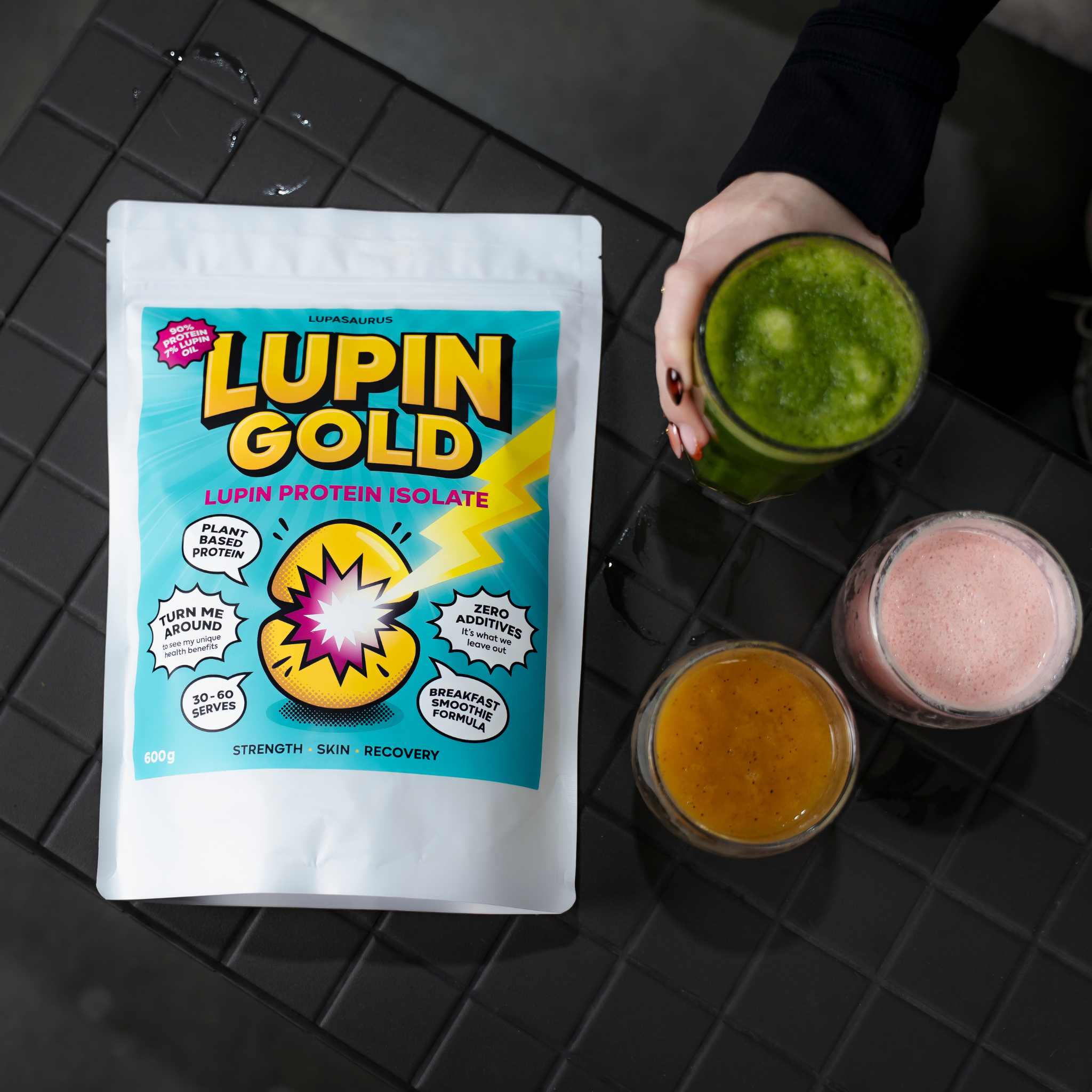 A pouch of Lupin Gold lupin protein isolate on a dark tiled bench beside three blended smoothies in green, pink and orange
