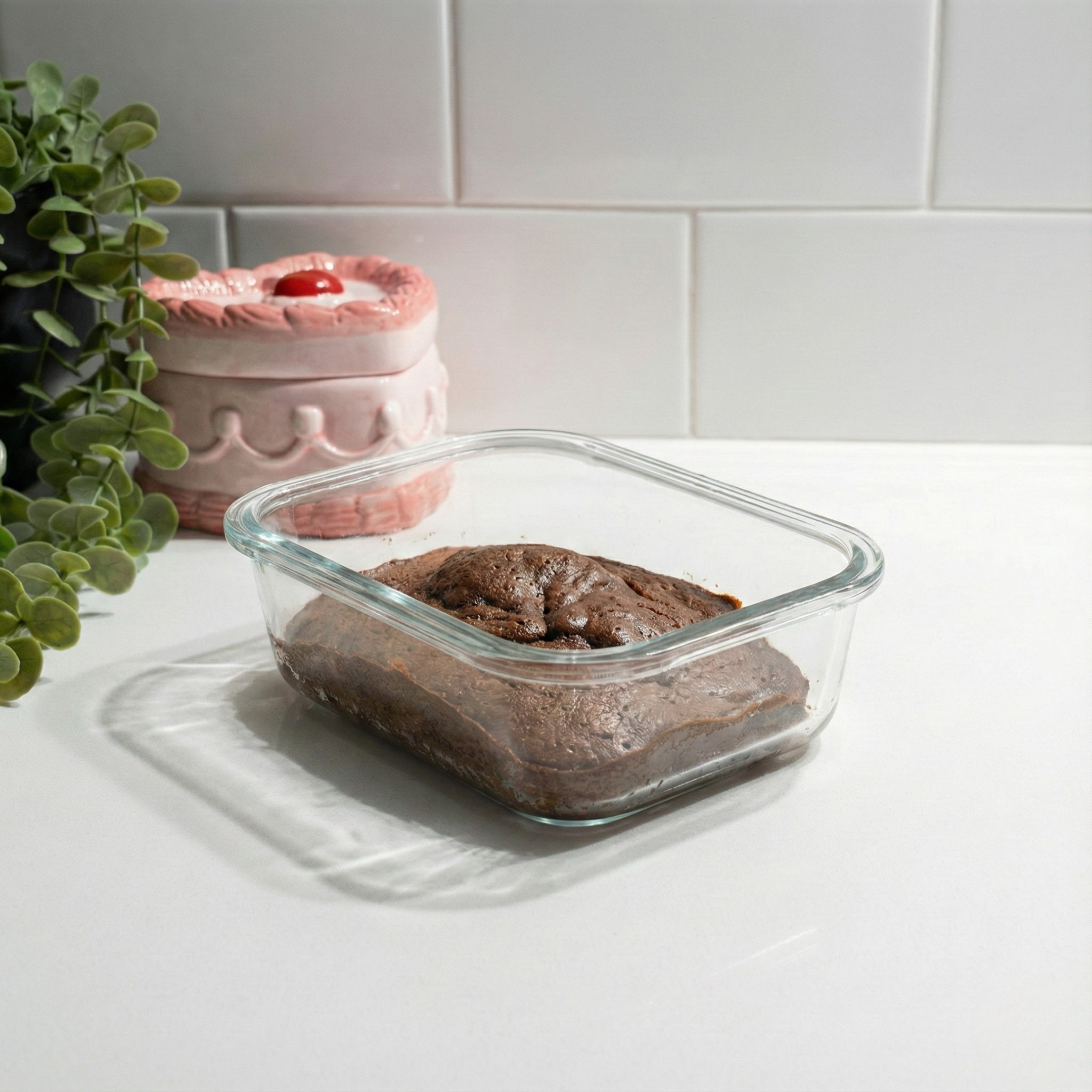 Glass dish of chocolate protein brownie mixture on a white kitchen bench beside a pink canister and a potted plant