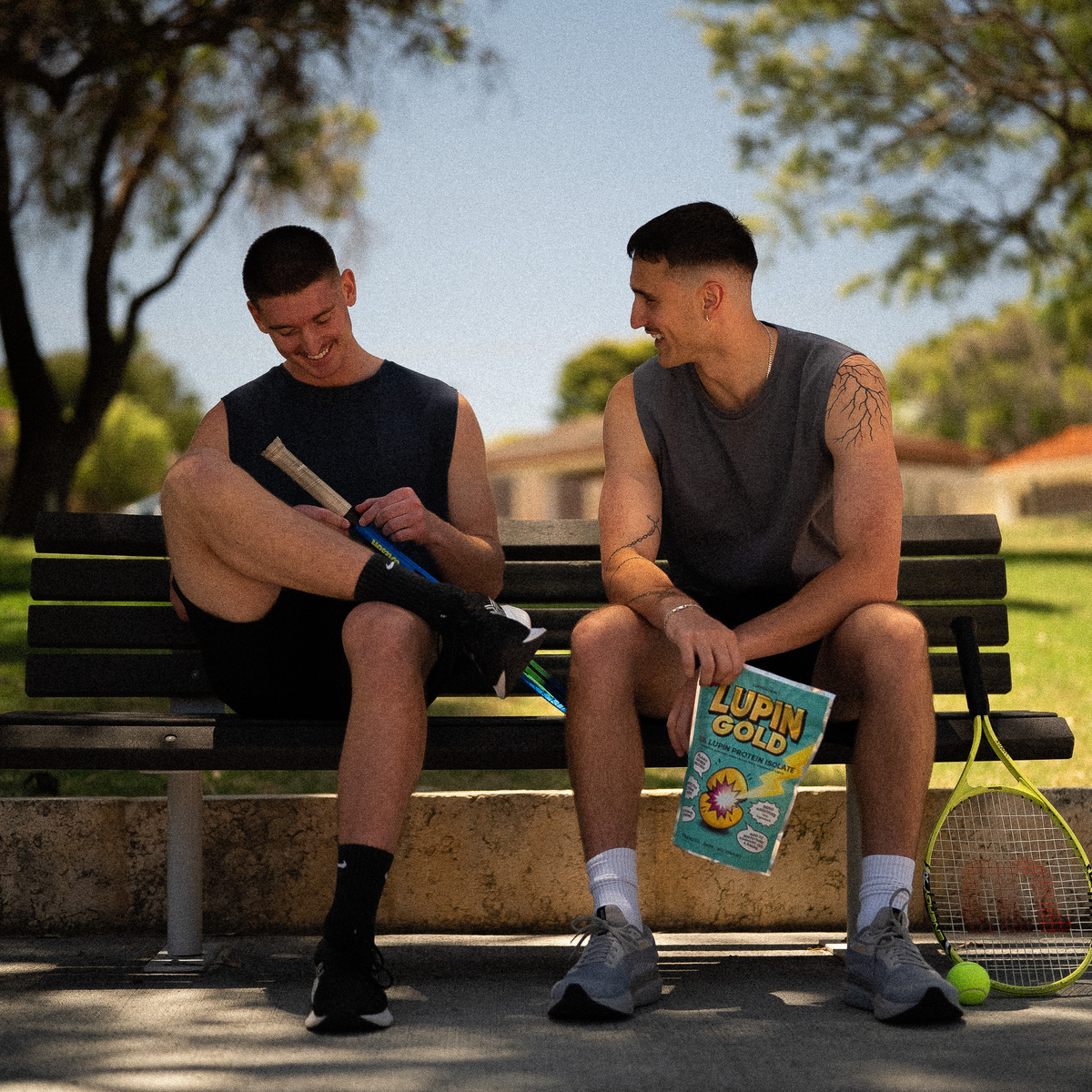 Two men in singlets sitting on a park bench with tennis rackets, one holding a pack of Lupin Gold lupin protein isolate