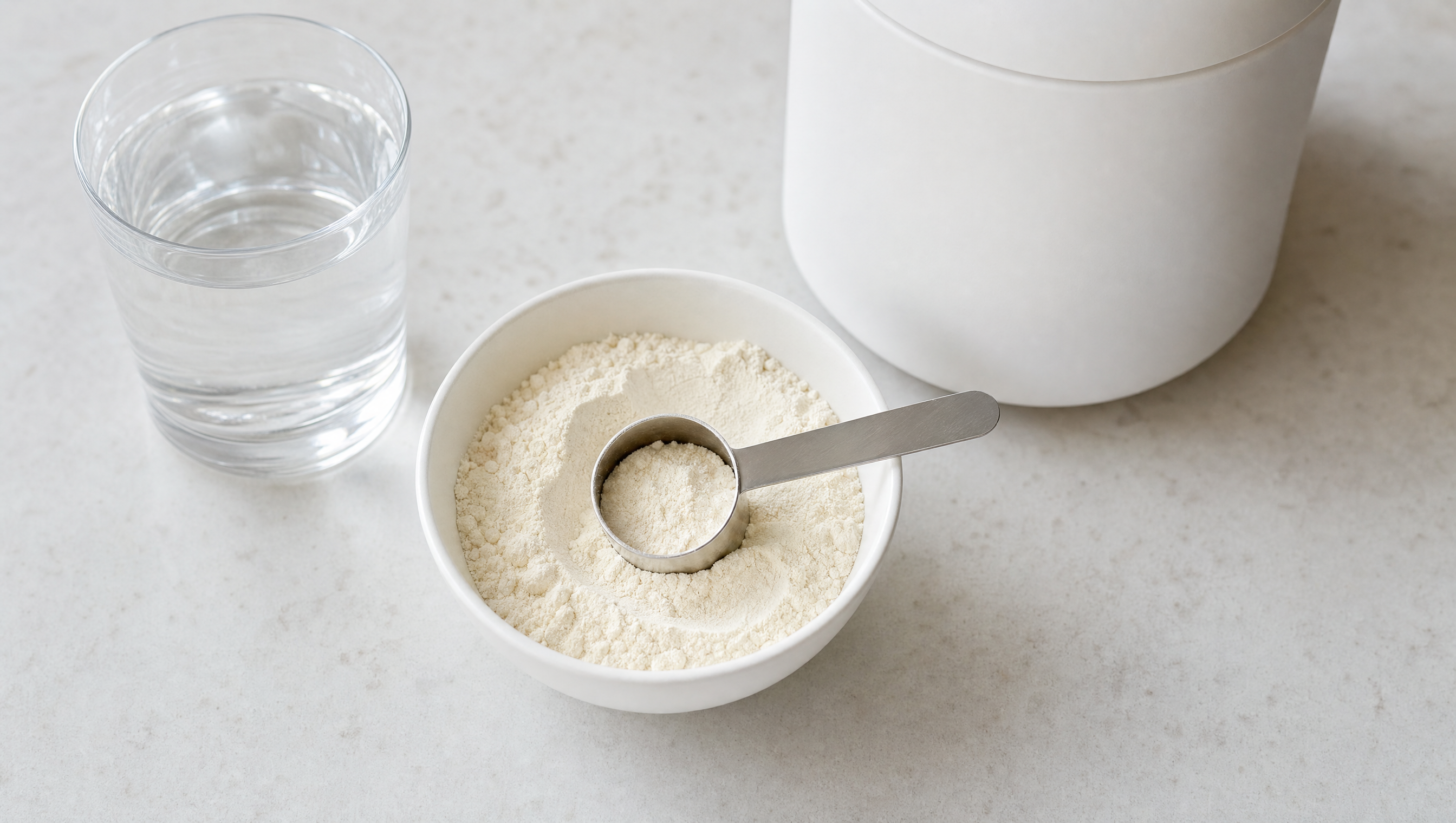 A white bowl of unflavoured protein powder with a stainless measuring scoop, a glass of water and a plain unbranded tub on a pale stone benchtop
