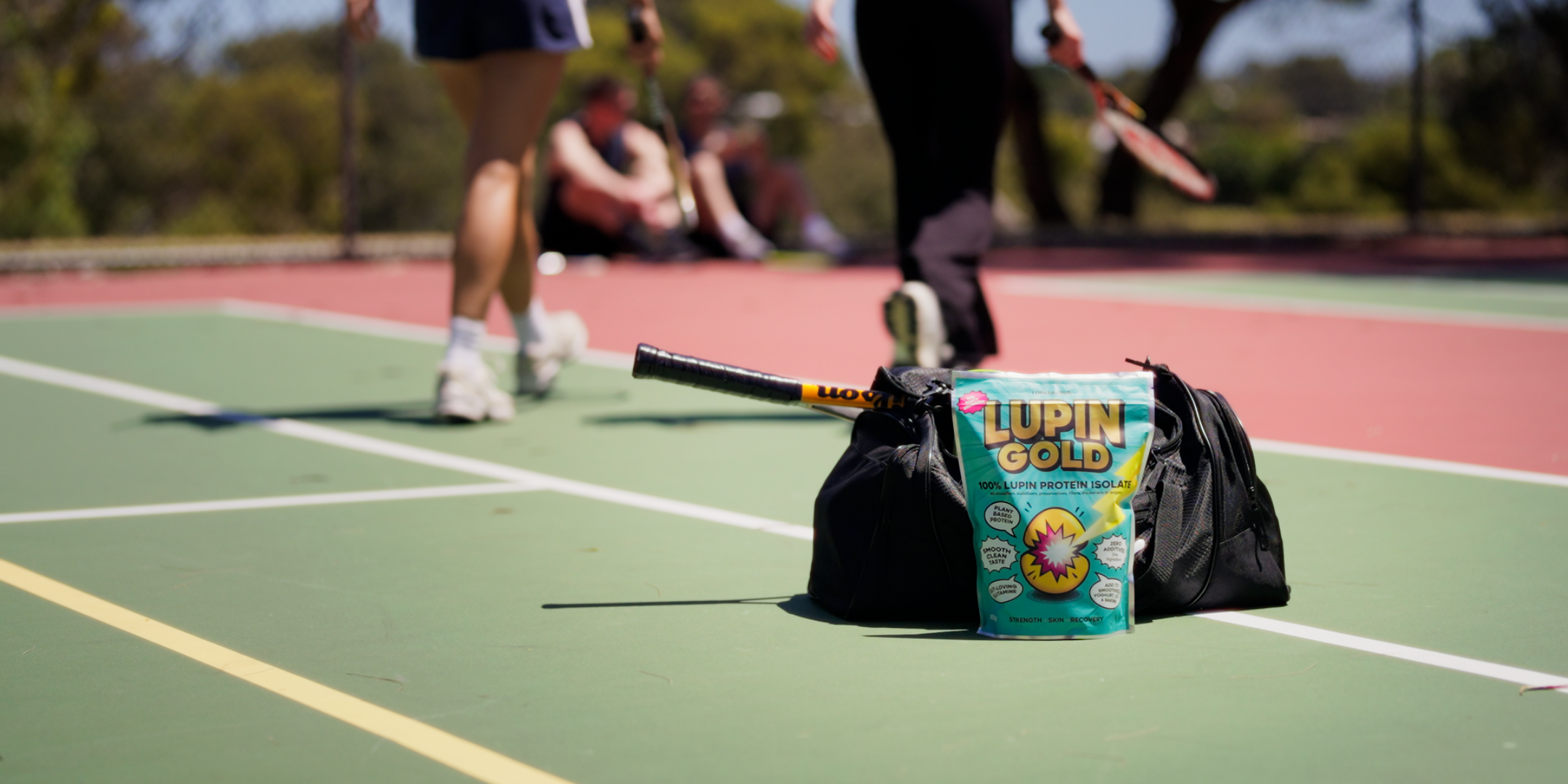 Woman in a cap and activewear sitting on a tennis court beside a gym bag holding a Lupin Gold pack, with tennis rackets and a ball