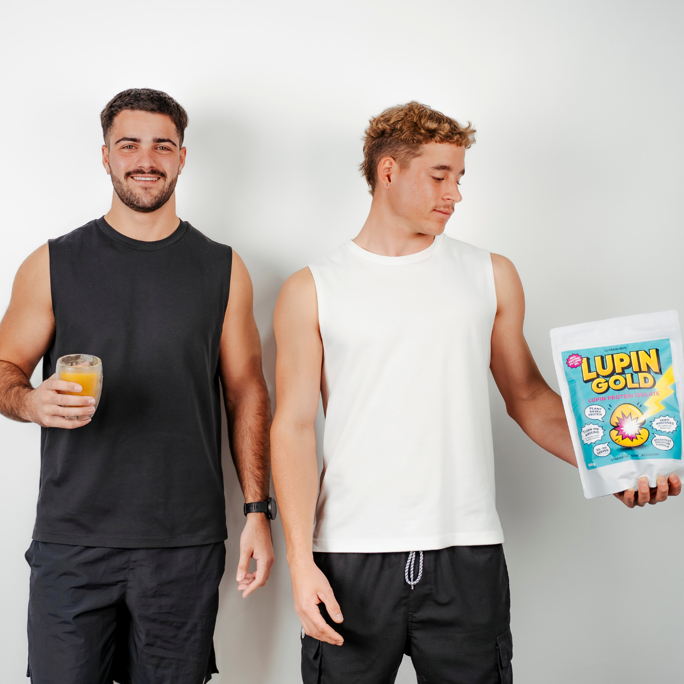 Two men standing side by side, one holding a glass of mango and lupin protein in a smoothie and the other holding a Lupin Gold protein isolate packet.