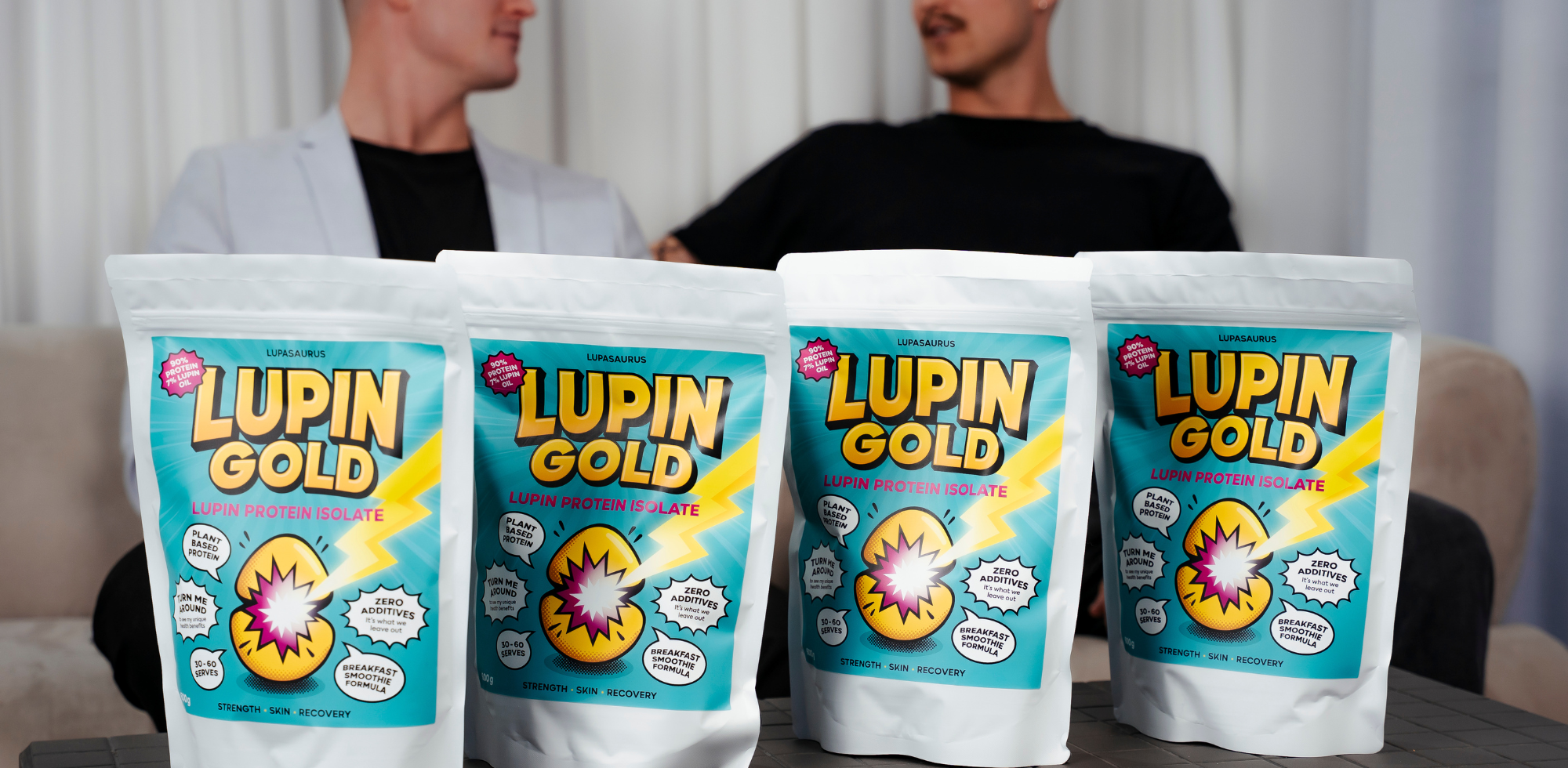 Four Lupin Gold protein powder packets on a table with two people in the background.