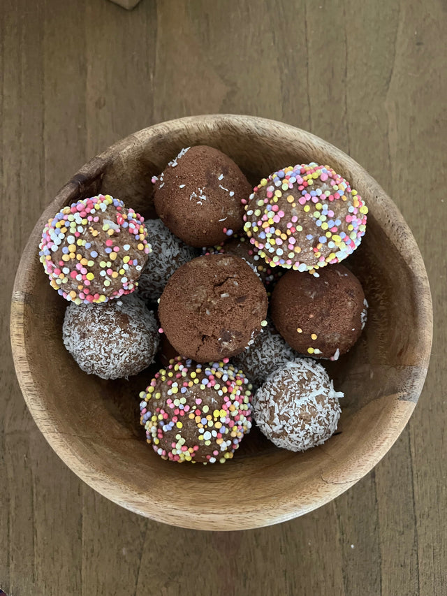 Wooden bowl of protein balls, some rolled in rainbow sprinkles, some in desiccated coconut and some plain cocoa
