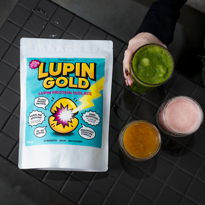 A pouch of Lupin Gold lupin protein isolate on a dark tiled bench beside three blended smoothies in green, pink and orange