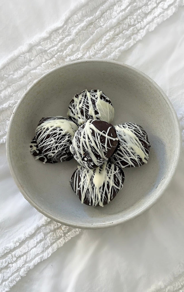 Five dark chocolate coated protein balls drizzled with white chocolate in a ceramic bowl on white linen