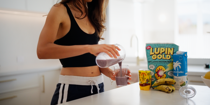 Woman in a black crop top pouring a purple protein smoothie into a glass in a white kitchen, with a Lupin Gold pack, bananas and oat milk on the bench