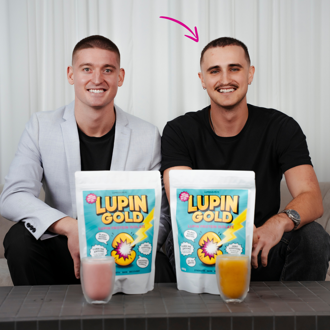 Kobi and Harvie (arrow pointing to his head) sitting behind Lupin Gold protein powder packaging with drinks, against a white curtain background.
