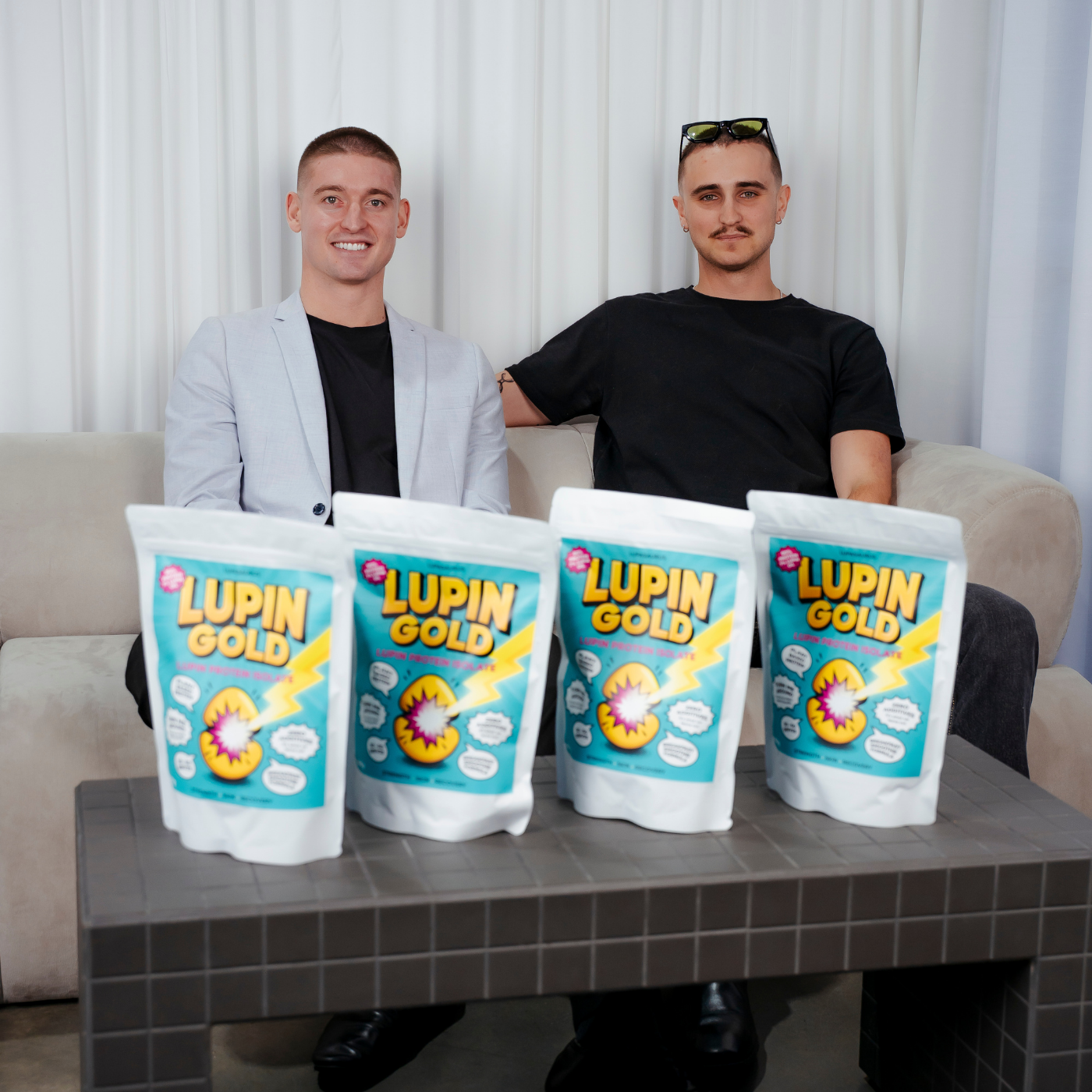 Kobi and Harvie sitting on a couch with four Lupin Gold bags in front of them.