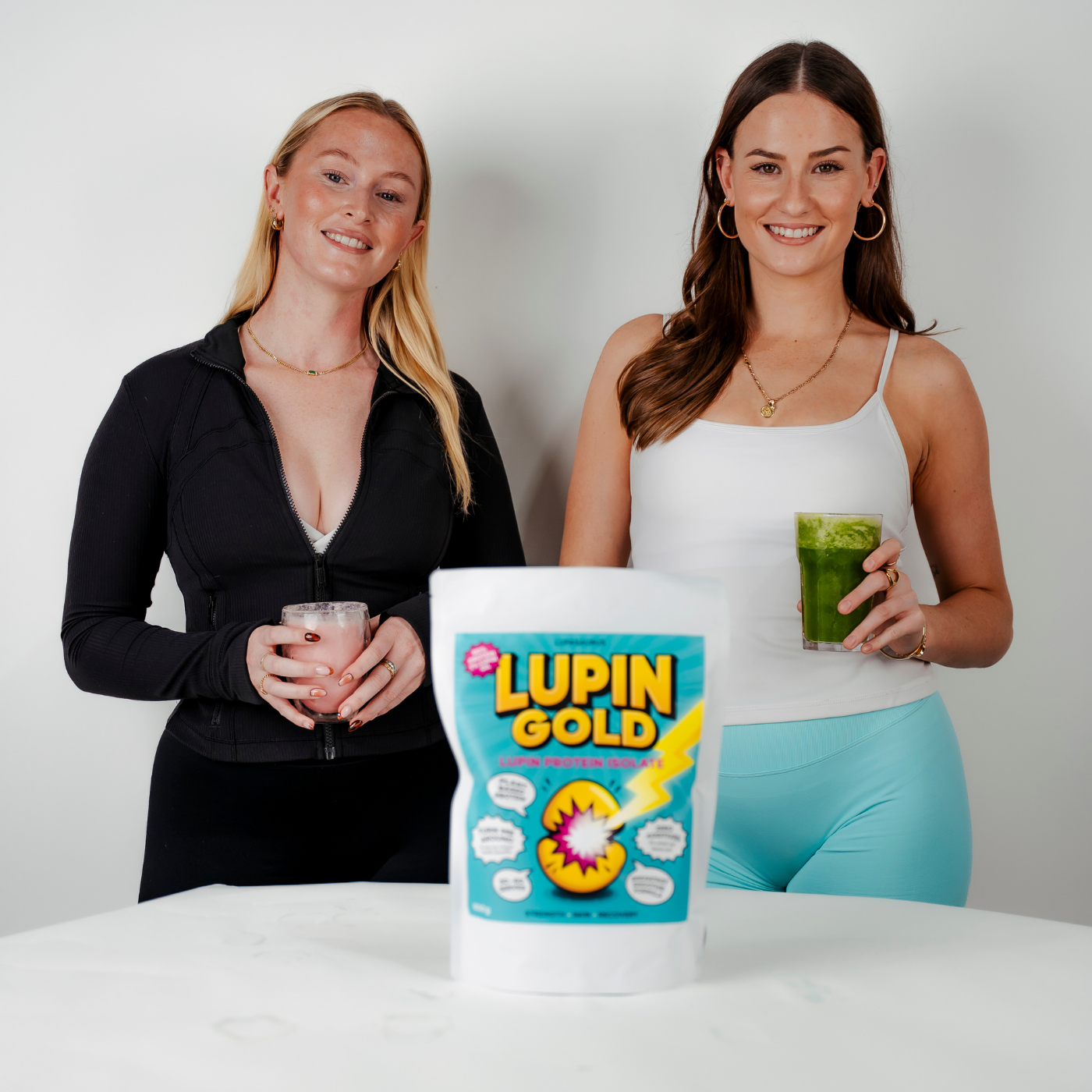 Two women holding Lupin protein smoothies with a Lupin Gold packet between them on a white surface.