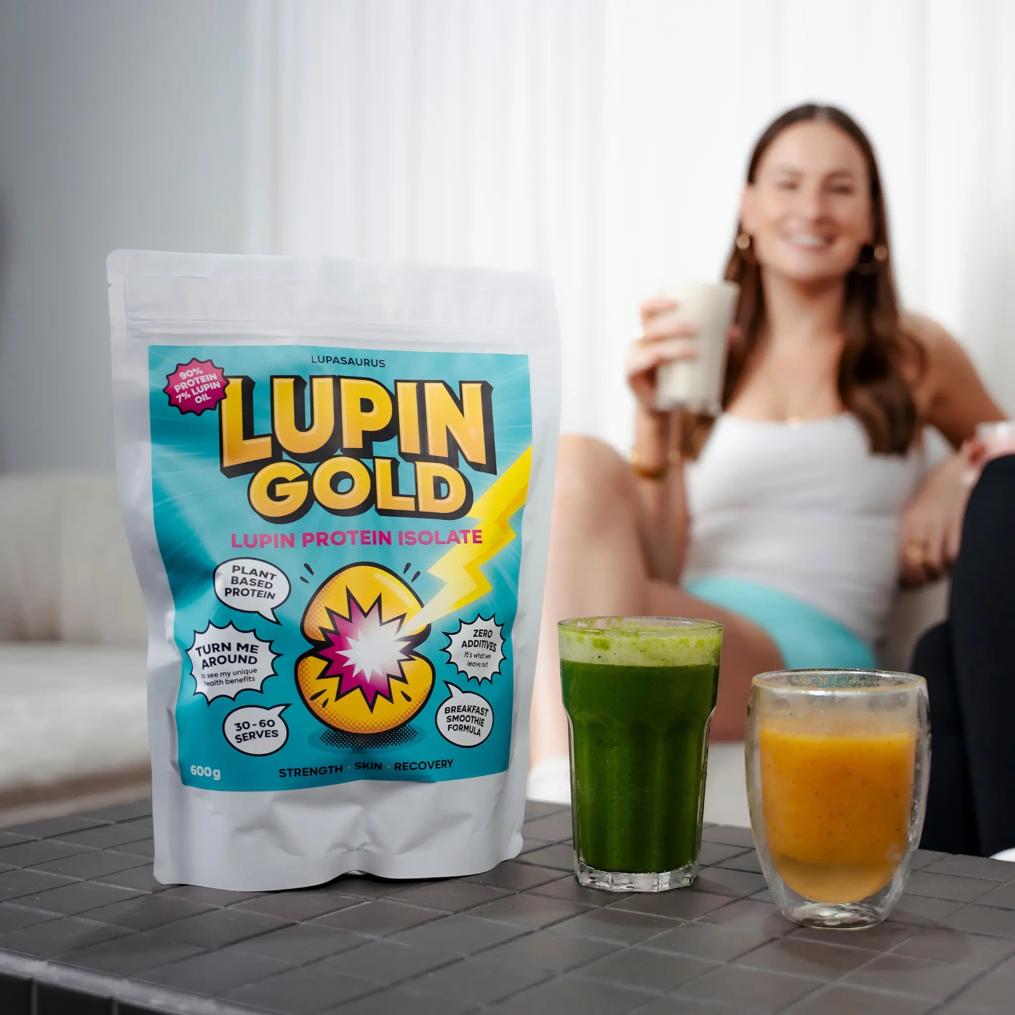 Lupin Gold protein package with a woman holding a glass of banana and lupin protein powder smoothie and two more smoothies on a table.