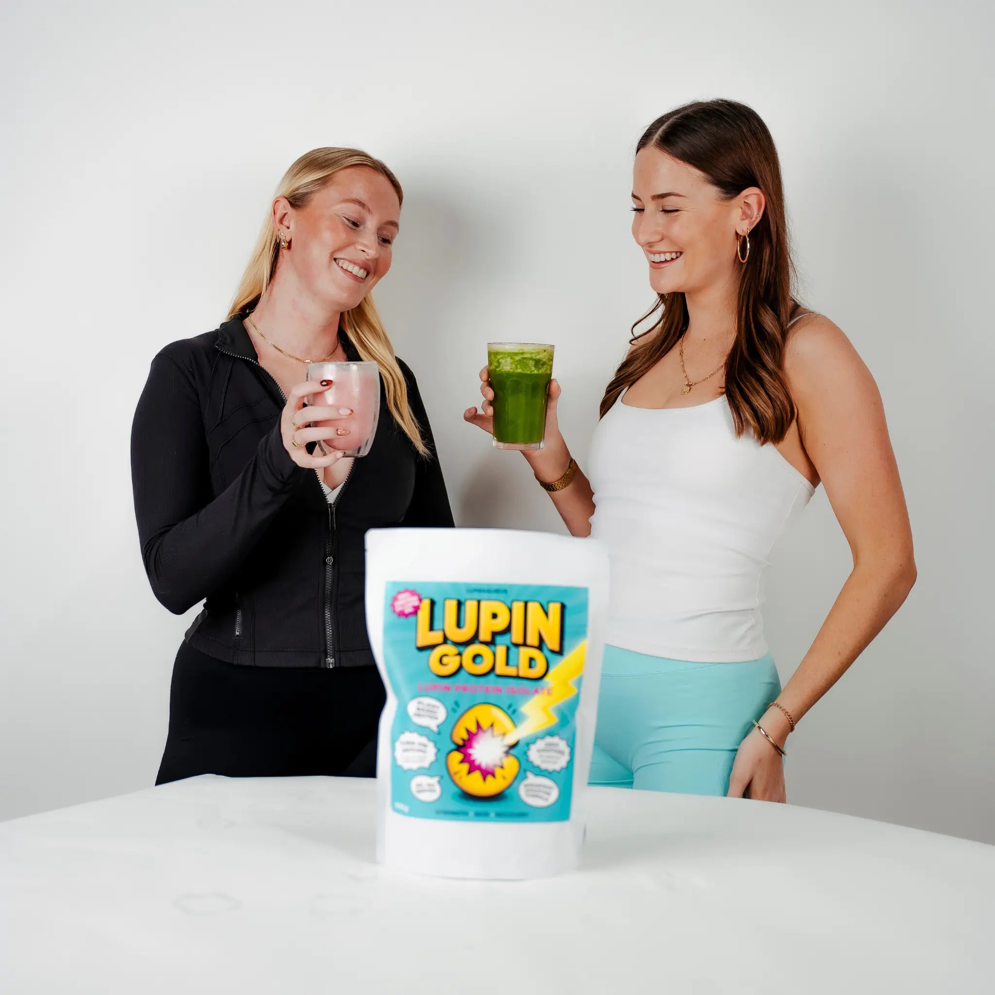 Two women holding drinks with a Lupin Gold protein powder packet in the foreground on a white background